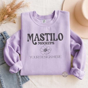Op de afbeelding: Een lichtpaarse sweatshirt met de tekst "MASTILO MOCKUPS" en "YOUR DESIGN HERE" in donkergrijs. Een bloemengrafiek staat onder de tekst. Een spijkerbroek en een beige hoed zijn op de achtergrond.