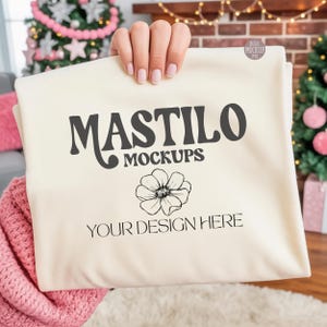 Puede incluir: Tela color crema con el texto "MASTILO MOCKUPS" y "YOUR DESIGN HERE" en negro. Un gráfico floral está debajo del texto. La tela está sostenida por una persona, con un árbol de Navidad y decoraciones en el fondo.
