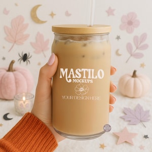 May include: A clear glass tumbler filled with a light brown beverage, topped with a wooden lid and a straw. The tumbler has the text "MASTILO MOCKUPS" and "YOUR DESIGN HERE" printed on it. The background features fall-themed decorations.