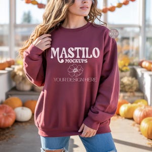 May include: A maroon sweatshirt with the text "MASTILO MOCKUPS" in white, along with a floral graphic and the words "YOUR DESIGN HERE". The sweatshirt is being worn by a person in a fall-themed setting with pumpkins.
