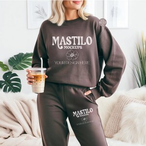 May include: A brown sweatshirt and sweatpants set with the text "MASTILO MOCKUPS" and a floral design. The model is holding a clear plastic cup with a straw filled with a layered beverage.