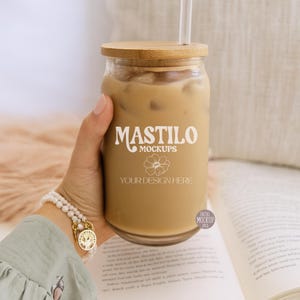 May include: A clear glass tumbler filled with iced coffee, topped with a wooden lid and a white straw. The tumbler has the text "MASTILO MOCKUPS" and "YOUR DESIGN HERE" printed on it. The drink is held in a hand.