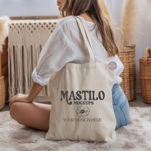 May include: A natural-coloured tote bag featuring the text "MASTILO MOCKUPS" and "YOUR DESIGN HERE" in black. The bag has long straps and is held by a person. The background includes baskets and decorative elements.