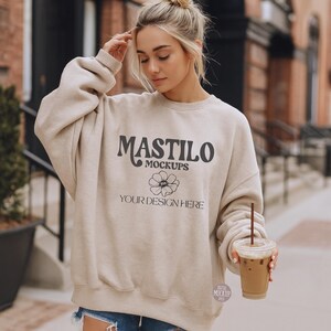 Könnte beinhalten: Beiges Sweatshirt mit dem Text "MASTILO MOCKUPS" und einem floralen Design. Die Worte "YOUR DESIGN HERE" stehen unter dem floralen Design. Das Sweatshirt ist übergroß und hat lange Ärmel.