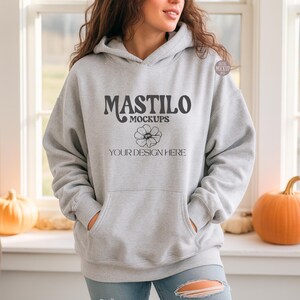 May include: A light gray hoodie with a front pocket and a hood. The hoodie features the text "MASTILO MOCKUPS" above a flower graphic, with "YOUR DESIGN HERE" below. The model is wearing blue jeans.