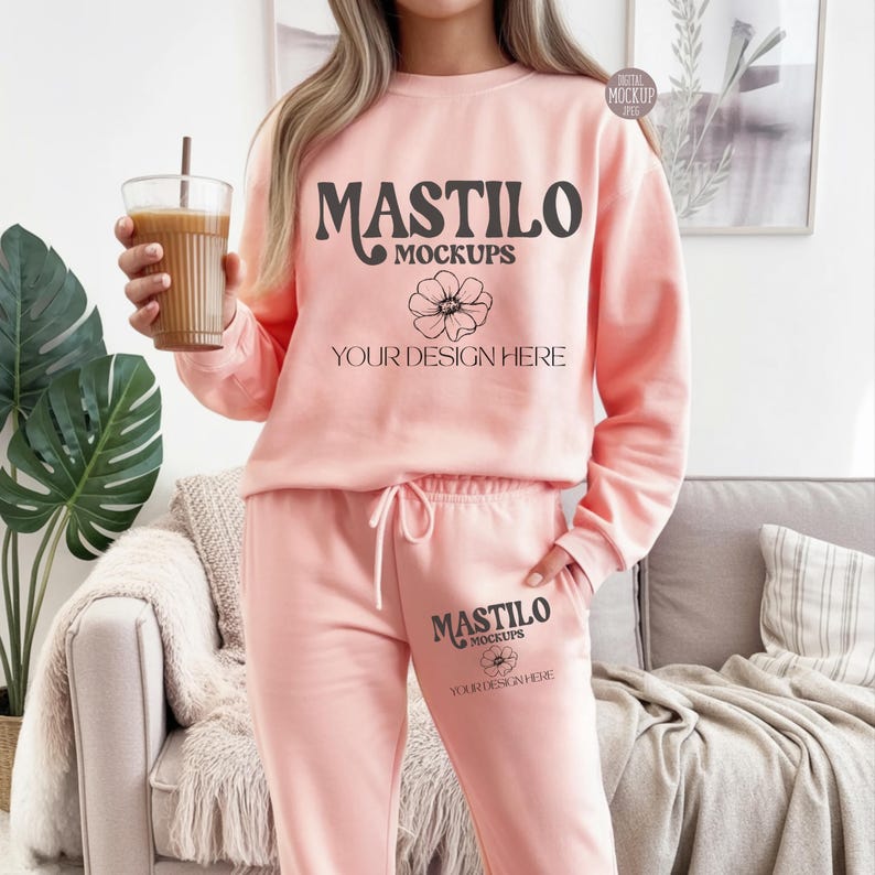 May include: A light pink sweatshirt and sweatpants set. The sweatshirt and sweatpants have the text "MASTILO MOCKUPS" and "YOUR DESIGN HERE" printed on them. The model is holding a glass of iced coffee.