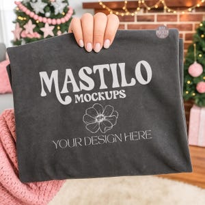 Puede incluir: Maqueta de camiseta gris oscuro con el texto "MASTILO MOCKUPS" y "YOUR DESIGN HERE" en blanco. Un gráfico floral está debajo del texto. La camiseta está sostenida por una persona.