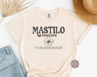 May include: Cream-colored t-shirt mockup with the text "MASTILO MOCKUPS" and "YOUR DESIGN HERE" in dark gray. A black floral graphic is below the text. A straw hat and blue jeans are partially visible.