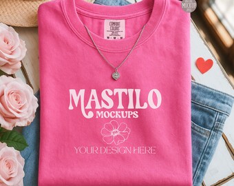 May include: A folded pink t-shirt with the text "MASTILO MOCKUPS" and "YOUR DESIGN HERE" in white. A silver necklace with a heart pendant rests on the shirt. The shirt is on top of blue jeans, with a straw hat and roses in the background.