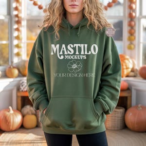 May include: An olive green hoodie with a front pocket and a drawstring hood. The hoodie features the text "MASTILO MOCKUPS" and "YOUR DESIGN HERE" in white. The background includes pumpkins and a wooden surface.