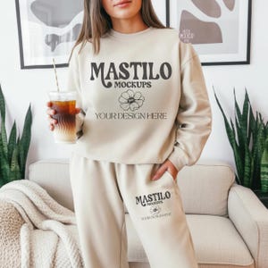 May include: A beige sweatshirt and sweatpants set with the text "MASTILO MOCKUPS" and a floral design. The set includes a sweatshirt and sweatpants. The person is holding a clear plastic cup with a straw.