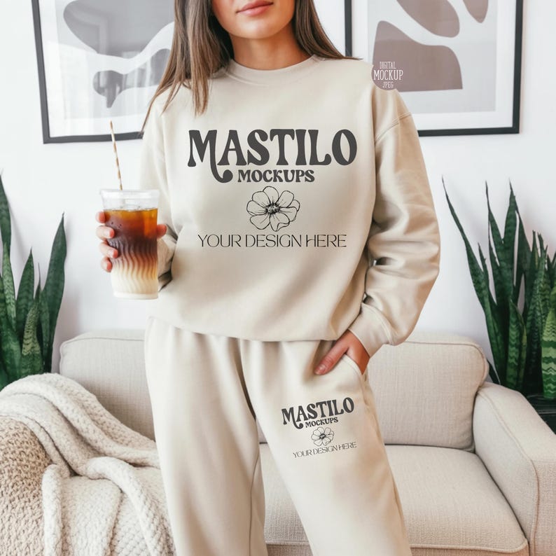 May include: A beige sweatshirt and sweatpants set with the text "MASTILO MOCKUPS" and a floral design. The set includes a sweatshirt and sweatpants. The person is holding a clear plastic cup with a straw.