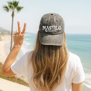 Könnte beinhalten: Graue, ausgewaschene Baseballkappe mit dem weißen Text "MASTILO MOCKUPS". Die Kappe wird von einer Person mit langen blonden Haaren getragen, vor dem Hintergrund des Ozeans und einer Palme. Die Person zeigt das Friedenszeichen.