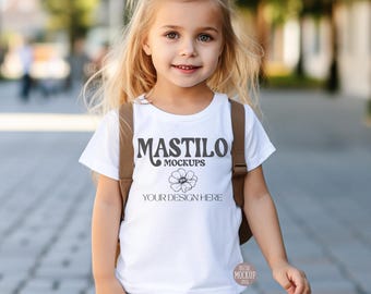 May include: A young girl wearing a white t-shirt with the text "MASTILO MOCKUPS" and a floral design. The shirt also includes the text "YOUR DESIGN HERE". She is wearing a brown backpack and light blue jeans.