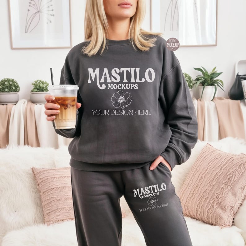 May include: Charcoal gray sweatshirt and sweatpants set with the text "MASTILO MOCKUPS" and "YOUR DESIGN HERE" in white. The person is holding a clear plastic cup with iced coffee. The set is a mockup for apparel design.