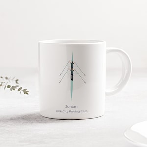 Peut inclure: Mug en céramique blanche avec une illustration turquoise et noire d'un bateau d'aviron. Le mug porte le nom "Jordan" et "York City Rowing Club" imprimé en dessous.