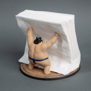 Op de afbeelding: Een novelty servethouder met een sumo worstelaar beeldje dat een stapel witte papieren servetten optilt. Het beeldje is bruin met een zwarte knot en een blauwe lendendoek. De basis is rond en van hout.