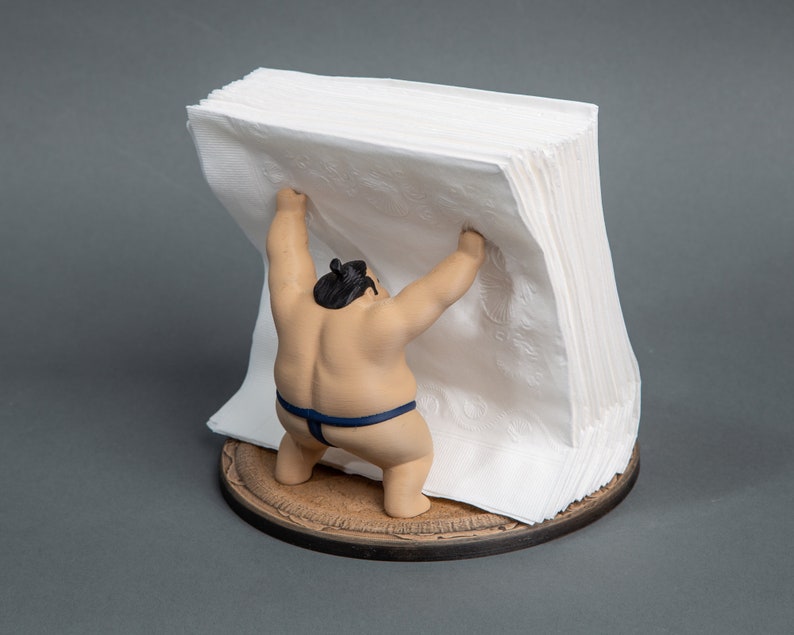Op de afbeelding: Een novelty servethouder met een sumo worstelaar beeldje dat een stapel witte papieren servetten optilt. Het beeldje is bruin met een zwarte knot en een blauwe lendendoek. De basis is rond en van hout.