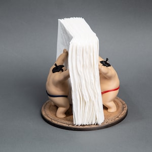 Op de afbeelding: Een novelty tissuehouder met twee sumo worstelaar beeldjes die een stapel witte tissues ondersteunen. De worstelaars dragen traditionele mawashi, &eacute;&eacute;n blauw, &eacute;&eacute;n rood. De basis is een rond houten platform.