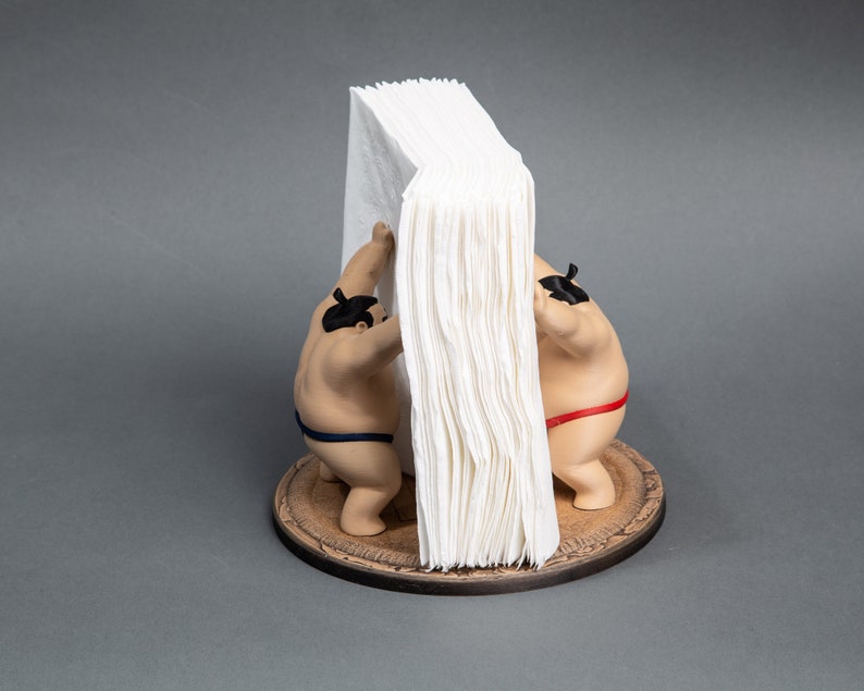Op de afbeelding: Een novelty tissuehouder met twee sumo worstelaar beeldjes die een stapel witte tissues ondersteunen. De worstelaars dragen traditionele mawashi, &eacute;&eacute;n blauw, &eacute;&eacute;n rood. De basis is een rond houten platform.
