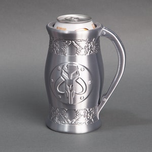 K&ouml;nnte beinhalten: Ein silberner Metall-Getr&auml;nkedosenhalter mit einem Mandalorianer-Symbol und filigranen Details. Der Halter ist f&uuml;r eine Getr&auml;nkedose in Standardgr&ouml;&szlig;e ausgelegt.