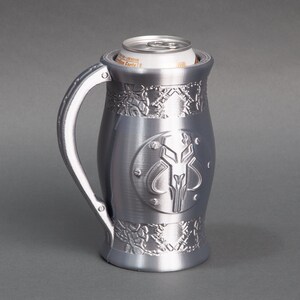 K&ouml;nnte beinhalten: Ein silberner Metall-Getr&auml;nkedosenhalter mit einem Mandalorianer-Symbol und filigranen Designs. Der Halter ist so konzipiert, dass eine Getr&auml;nkedose kalt bleibt.