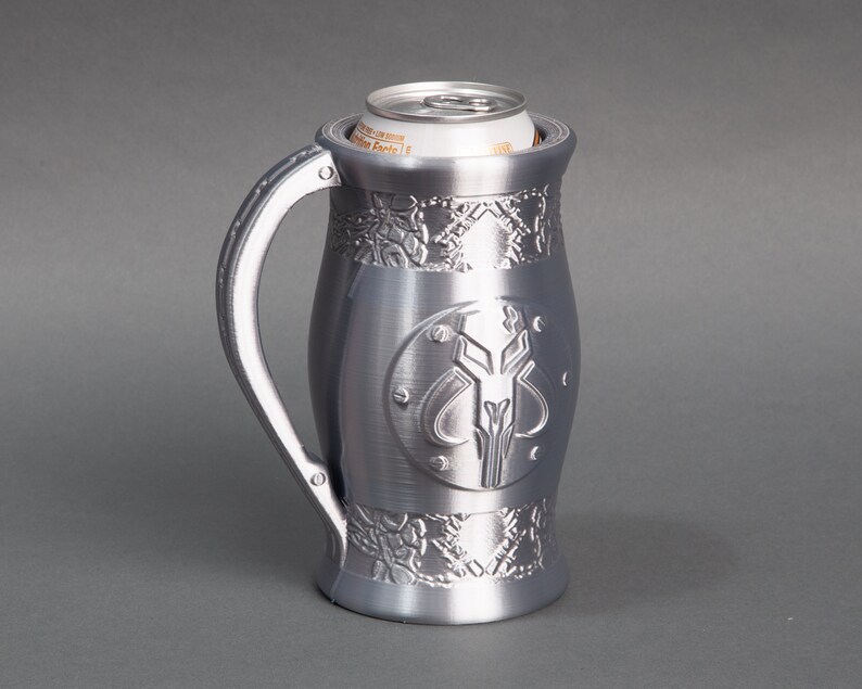 K&ouml;nnte beinhalten: Ein silberner Metall-Getr&auml;nkedosenhalter mit einem Mandalorianer-Symbol und filigranen Designs. Der Halter ist so konzipiert, dass eine Getr&auml;nkedose kalt bleibt.