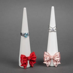 May include: Two white cone-shaped ring displays. One holds a silver ring with a large blue gemstone, the other a silver criss-cross ring. Each display has a decorative bow at the base; one red, one pink.