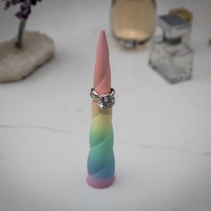 May include: A pastel-colored unicorn horn ring holder with a silver ring featuring a large, clear gemstone. The horn transitions from pink to yellow, green, and blue. The ring is displayed on the top of the horn, showcasing its design.