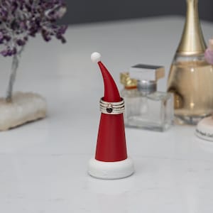 May include: A red and white Santa hat-shaped ring holder with rings on it. The ring holder has a white base and a white pom-pom at the tip. The rings are silver with a dark red stone. Other items are in the background.