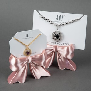 May include: Two necklaces displayed on white cards with pink satin bows. One necklace features a gold chain with a round purple stone pendant. The other is a silver chain with a round black stone pendant featuring star and moon engravings.