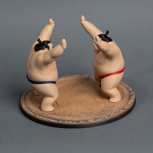 Op de afbeelding: Twee sumo worstelaar beeldjes in een vechthouding, elk met een zwarte knot en lendendoek. E&eacute;n worstelaar draagt een blauwe lendendoek, de andere een rode. Ze staan op een ronde, getextureerde, bruine basis.