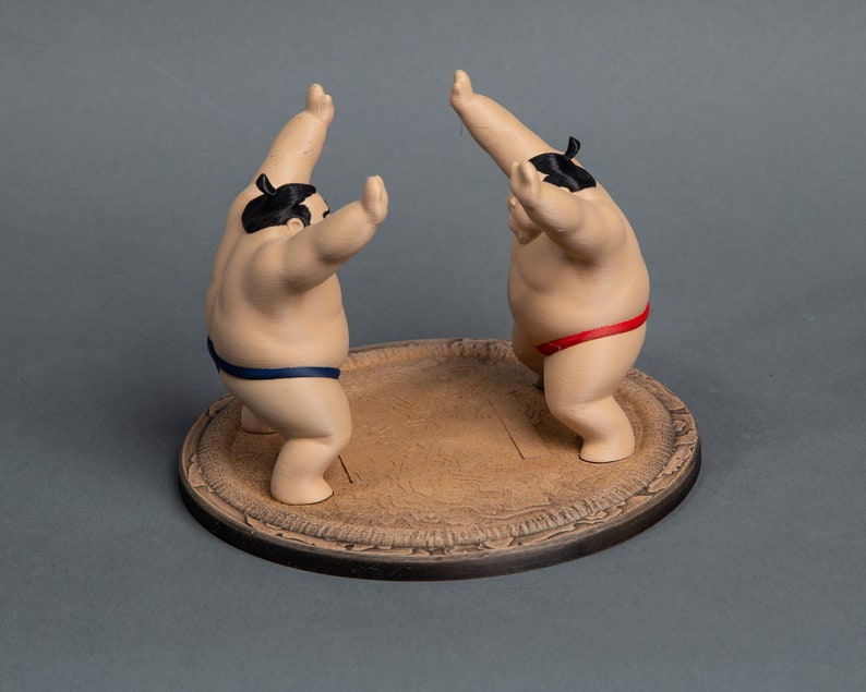 Op de afbeelding: Twee sumo worstelaar beeldjes in een vechthouding, elk met een zwarte knot en lendendoek. E&eacute;n worstelaar draagt een blauwe lendendoek, de andere een rode. Ze staan op een ronde, getextureerde, bruine basis.