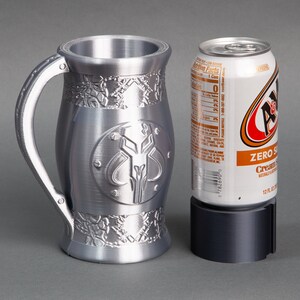 K&ouml;nnte beinhalten: Eine silberne Metalltasse mit einem Mandalorianer-Symbol und filigranen Details. Die Tasse steht neben einer Dose A&W Cream Soda Zero Sugar.