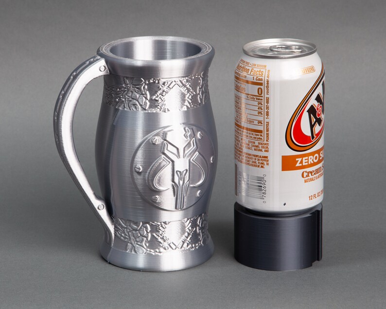 K&ouml;nnte beinhalten: Eine silberne Metalltasse mit einem Mandalorianer-Symbol und filigranen Details. Die Tasse steht neben einer Dose A&W Cream Soda Zero Sugar.