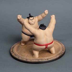 Op de afbeelding: Een beeldje van twee sumoworstelaars in een wedstrijd. De beige worstelaars dragen zwarte knotjes en blauwe en rode mawashi. Ze staan op een bruine, cirkelvormige basis met een sierlijke rand.