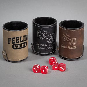 May include: Three brown leather dice cups with different engraved designs. The cups are on a gray surface with five red dice scattered around them. The designs on the cups are "Feeling Lucky", "Oxford Grand Senior Living", and "Let's Roll!"