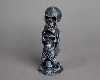 May include: A silver sculpture of three stacked skulls, each with hands covering their eyes, ears, and mouth. The sculpture is on a base with bone-like details.