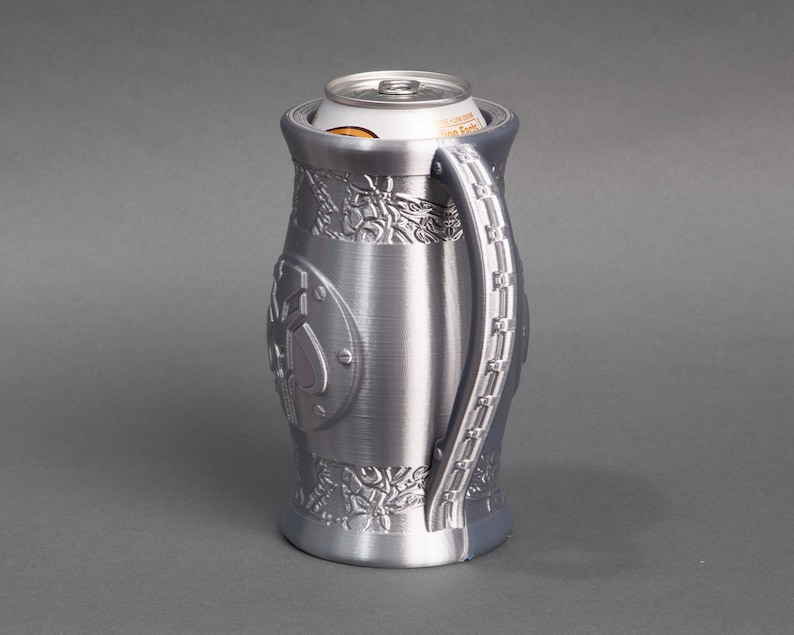 K&ouml;nnte beinhalten: Ein silberner Metall-Getr&auml;nkedosenhalter mit einem komplizierten Design und einem Griff. Der Halter ist f&uuml;r eine Standard-Getr&auml;nkedose ausgelegt.