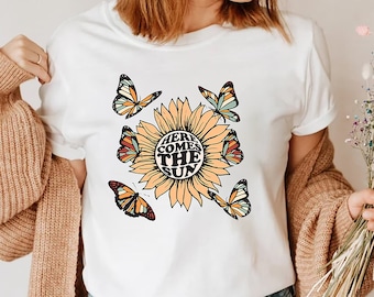 May include: White t-shirt with a sunflower graphic and the text "Here Comes The Sun". The sunflower has colorful butterflies around it.