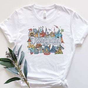 Puede incluir: Camiseta blanca con una ilustraci&oacute;n colorida de Disneyland Resort, incluyendo monumentos emblem&aacute;ticos como el Castillo de la Bella Durmiente, Space Mountain y el Matterhorn. El texto "Disneyland Resort" est&aacute; impreso debajo de la ilustraci&oacute;n.