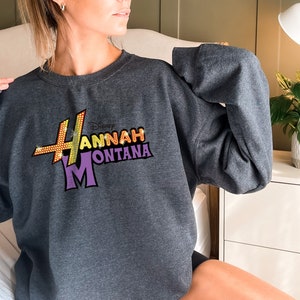 Puede incluir: Una sudadera gris con un gr&aacute;fico brillante de color morado, amarillo y naranja del programa de Disney Channel "Hannah Montana".