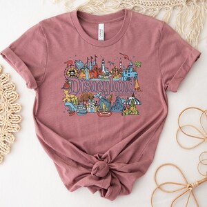 Puede incluir: Una camiseta de color malva con un estampado gr&aacute;fico de Disneyland Resort. El gr&aacute;fico presenta una ilustraci&oacute;n colorida de varias atracciones de Disneyland, incluyendo el Castillo de la Bella Durmiente, el Matterhorn y la Mansi&oacute;n Encantada.