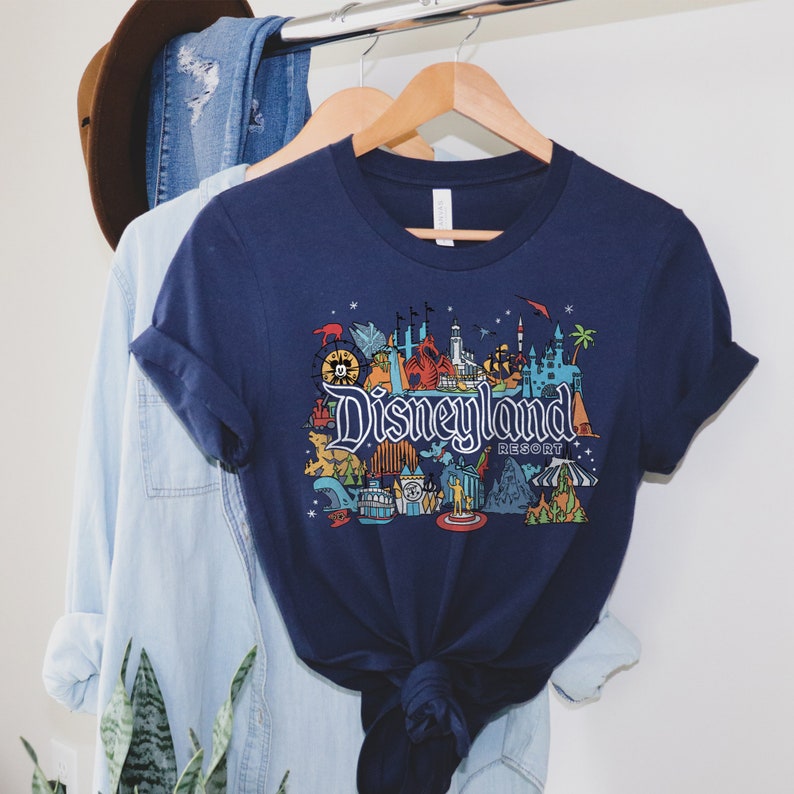 Puede incluir: Una camiseta azul marino con un gr&aacute;fico colorido de iconos de Disneyland Resort, incluyendo el Castillo de la Bella Durmiente, Space Mountain y el Matterhorn. El texto "Disneyland Resort" est&aacute; impreso en blanco.