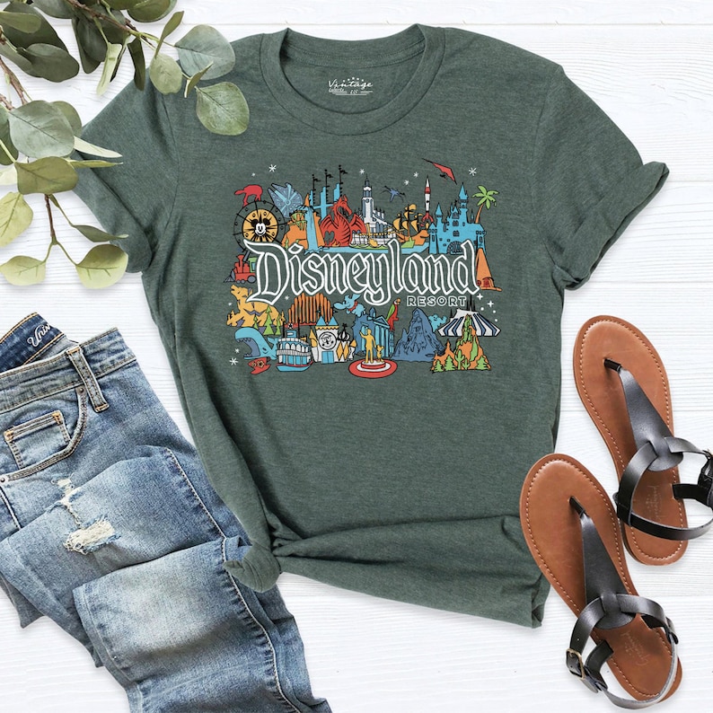 Puede incluir: Una camiseta verde con un gr&aacute;fico colorido de iconos de Disneyland Resort, incluyendo el castillo, los paseos y los personajes. El texto "Disneyland Resort" est&aacute; impreso en la camiseta.