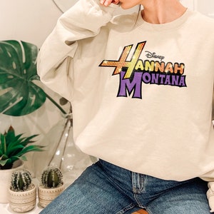 Puede incluir: Una sudadera beige con un estampado gr&aacute;fico morado y amarillo que dice "Disney Hannah Montana".