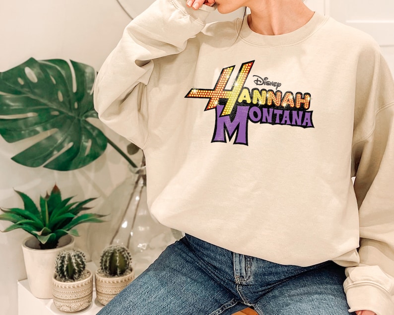 Puede incluir: Una sudadera beige con un estampado gr&aacute;fico morado y amarillo que dice "Disney Hannah Montana".