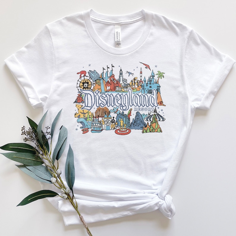 Puede incluir: Camiseta blanca con una ilustraci&oacute;n colorida de Disneyland Resort, incluyendo monumentos emblem&aacute;ticos como el Castillo de la Bella Durmiente, Space Mountain y el Matterhorn. El texto "Disneyland Resort" est&aacute; impreso debajo de la ilustraci&oacute;n.