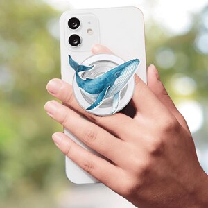 May include: A white smartphone featuring a circular collapsible phone grip attached to the back. The grip displays a detailed watercolour illustration of a blue whale with a white underbelly, swimming in a curved motion across the circular surface.