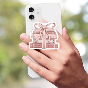 May include: A circular phone grip featuring a stack of four pink books tied with a matching ribbon. The book spines display the text "FORCED PROXIMITY," "FATED MATES," and "ENEMIES TO LOVERS" in a clear, dark font against the light pink background.